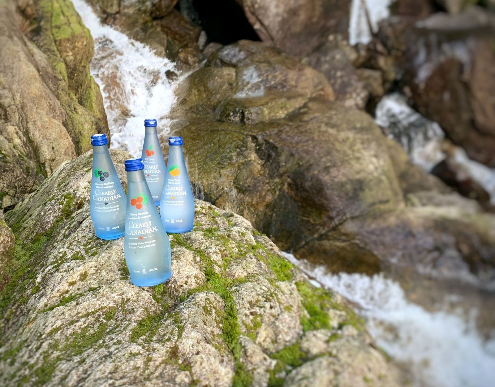 Clearly Canadian - Four 325ml Bottles of All Four Flavours On a Rock by a Mountain Stream